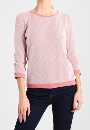 Hellrosa strukturierter Pullover mit rundem Ausschnitt und dreiviertel Ärmeln, mit einem kontrastierenden pinken Saum und Bündchen. Kombiniert mit dunklen Jeans.