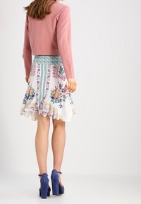 Pull court rose sur une jupe blanche ornée de motifs floraux colorés, taille smockée et escarpins bleus.