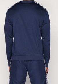 Námořnická modrá sportovní mikina s dlouhými rukávy, žebrovanými manžetami a pasem, se bílými postranními akcenty a hladkou texturou.