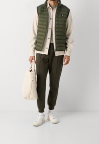 Grön pufferväst över en krämfärgad skjorta, med olivfärgade joggingsbyxor och vita sneakers. Bär en beige tote-väska. Släta texturer och avslappnad design.