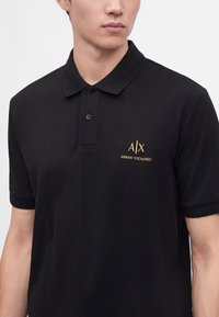 Jeune homme portant un polo noir avec un col et deux boutons, avec un petit logo doré Armani Exchange sur la poitrine gauche.
