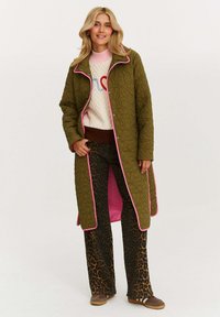 Prešívaný dlhý olivový kabát s ružovým lemovaním, nosený cez vzorovaný sveter a nohavice s leopardím vzorom, prezentovaný s bočnými vreckami a patentkami.