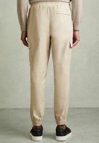 Pantalones cargo beige con cintura elástica, bolsillos laterales y puños elásticos; combinados con zapatillas oscuras sobre un fondo gris.