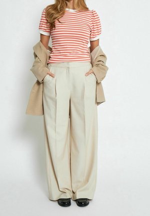 Vrouw draagt beige wijde broekspijpen, zwarte schoenen, een rood-wit gestreept T-shirt met korte mouwen en een beige jas over haar armen gedrapeerd.
