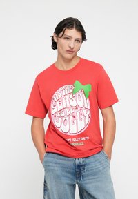 Crvena pamučna majica s rundastim grafičkim prikazom i bijelim natpisom "'Tis the season to be jolly" te zelenom mašnom kao akcentom. Ležeran kroj.