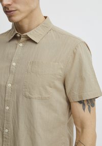 Camisa de botones de manga corta color beige, hecha de una tela ligera, que cuenta con un bolsillo en el pecho y un cuello clásico de punta.