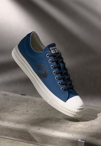 Blue canvas sneaker with a white rubber toe cap, black star logo, and dark gray laces. Features textured detailing on the sole.