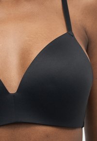 Κοντινό πλάνο ενός ατόμου που φοράει ένα λείο, μαύρο, αμάνικο σουτιέν με λεπτές τιράντες πάνω σε καφέ δέρμα.