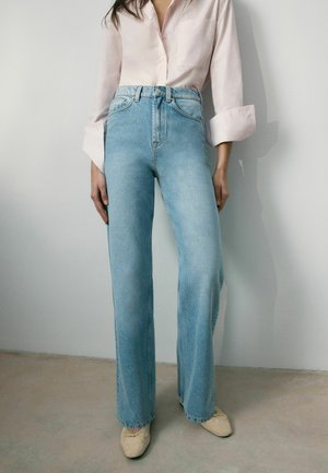 Person iført lyseblå, højtaljede, lige jeans, en lyserød skjorte med knapper og opkørte ærmer, samt beige ballet flats.