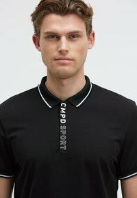 Polo negro con cuello y puños blancos en contraste, que presenta un logo vertical "CMPD SPORT" en la tapeta, fabricado con tejido suave.