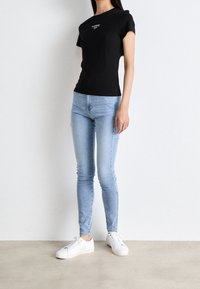 Camiseta negra de manga corta con logo blanco, combinada con jeans ajustados de color azul claro y zapatillas blancas, de pie frente a un fondo neutro.