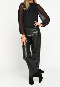 Blusa nera con maniche trasparenti a pois, abbinata a pantaloni larghi in finta pelle nera a vita alta e stivali neri alla caviglia.
