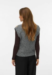 Suéter sin mangas gris con un acabado suave y texturizado, con un escote amplio y una camiseta de manga larga marrón contrastante debajo.