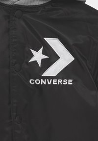 Giacca coach in satin nera con cappuccio grigio, bottoni a pressione e un logo ricamato bianco a forma di stella e freccia con il testo "CONVERSE".