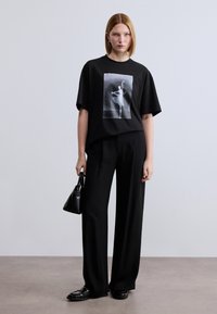 Μαύρο υπερμεγέθες t-shirt με φωτογραφικό γραφικό, σε συνδυασμό με μαύρο παντελόνι φαρδύ και μαύρα loafers, κρατώντας μια μικρή μαύρη τσάντα.