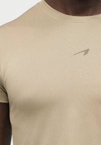 Μπεζ εφαρμοστό κοντομάνικο μπλουζάκι με μικρό, λοξό λογότυπο στο επάνω αριστερό μέρος του στήθους, που φοριέται από άτομο με σκούρο δέρμα.