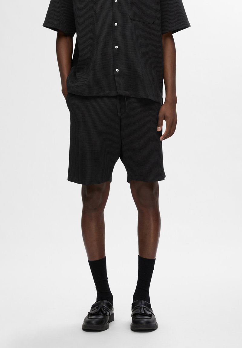 Fekete bordázott, rövidujjú póló zsebbel, hozzá illő fekete rövidnadrággal, amely rugalmas derékkal és húzózsinórral rendelkezik. Fekete cipő és zokni.