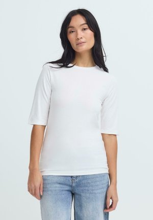 Vrouw met lang donker haar, gekleed in een wit shirt met korte mouwen en lichtblauwe jeans, staand tegen een effen lichte achtergrond.