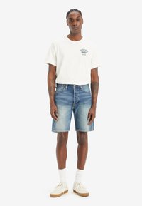 Maglietta bianca in cotone con stampa verde, abbinata a pantaloni corti in denim blu. Indossata con calzini bianchi e sneaker chiare, caratterizzate da un design testurizzato.