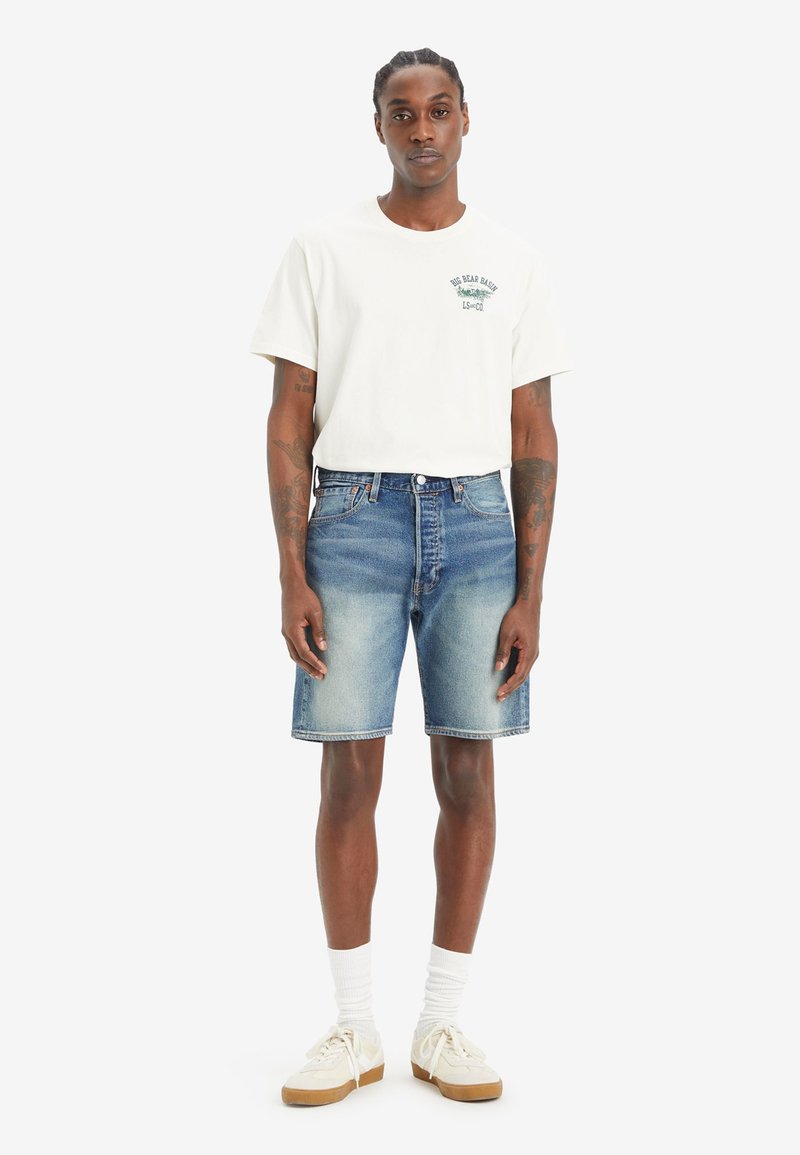 Weißes Baumwoll-T-Shirt mit grünem Druck, kombiniert mit blauen Jeansshorts. Getragen mit weißen Socken und hellen Sneakern mit strukturiertem Design.