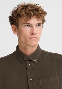 Camisa de botones de pana marrón con cuello, bolsillo frontal y botones marrones, con una tela texturizada y un diseño ajustado.