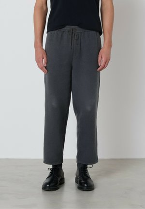 Eine Person trägt graue Kordelzughosen, schwarze Socken, schwarze Schnürschuhe und ein schwarzes Shirt und steht auf einem hellgrauen Boden.