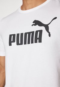 Camiseta blanca de manga corta con un logo negro de "PUMA" y un gráfico de un puma saltando en el pecho. Fabricada con un material suave y ligero.