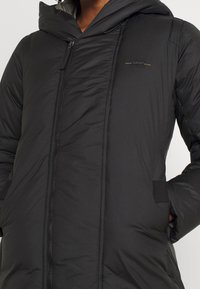 Černá péřová bunda s kapucí, disponující předním zipem, prošívanou strukturou a bočními kapsami. Výrazný detail loga v zlaté barvě.