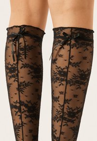 Schwarze Spitzen-Strümpfe bis zur Oberschenkelhöhe mit floralen Mustern, versehen mit einem gewellten Saum und schwarzen Schleifen, die den oberen Oberschenkel betonen.