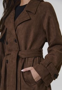 Kobieta w ciemno-brązowym zamszowym płaszczu z paskiem zawiązanym w pasie, z ręką w kieszeni, na czarnym topie.