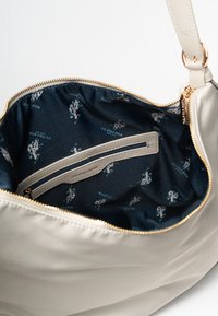 Beige handväska med dragkedjestängning, med ett mörkblått foder som pryds av upprepade U.S. Polo Assn. logotypmönster och en liten dragkedjeficka.