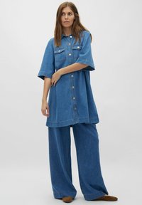 Denim jurkje in medium blauw, met korte mouwen, knopen aan de voorkant en twee borstzakken. Gecombineerd met ruimvallende denim broek.