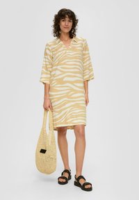 Kurzes ärmelloses Kleid in hellgelb mit weißen Zebra-Streifen, das über einen V-Ausschnitt verfügt. Das Modell hält eine gewebte beige Tasche und trägt schwarze Sandalen.