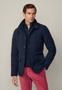 Giacca trapuntata blu navy con colletto alto, bottoni a scatto e tasche frontali, indossata sopra una giacca interna con zip, abbinata a pantaloni in velluto a coste rosa.