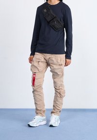 Osoba w beżowych spodniach cargo z czerwonym metką, białych i szarych sneakersach, granatowej koszuli z długim rękawem i czarnej torbie na ramię.