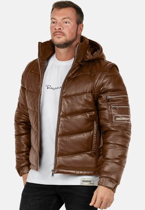 Man die een bruine gewatteerde leren jas met capuchon draagt over een wit t-shirt met grafische print en zwarte broek, staand tegen een effen achtergrond.