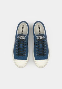 Blue canvas sneakers with white rubber toe caps, black laces, and contrasting side star logo. Text on the tongue reads "Converse All Star."