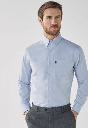 Hellblaues Langarm-Hemd mit Knopfverschluss und spitzem Kragen, einer einzelnen Brusttasche und einer glatten Textur. Kombiniert mit grauen Hosen.