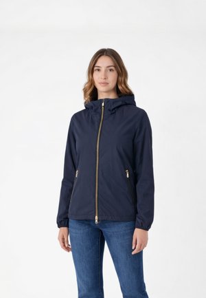 Donna che indossa una giacca blu navy con cappuccio, cerniera dorata e tasche, abbinata a jeans blu, in piedi su sfondo bianco.