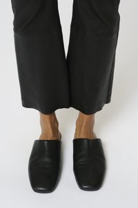 Sorte lædermules med firkantet tå og glat tekstur, parret med brede sorte bukser, der viser et minimalistisk design.