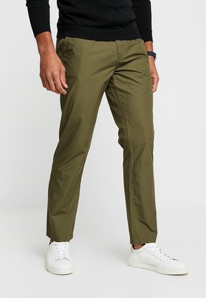 Homme portant un pantalon olive droit, une chemise noire à manches longues, des baskets blanches et une montre-bracelet sombre, debout sur fond blanc.