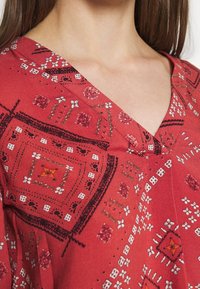 Blouse rouge à motifs avec un décolleté en V, présentant des motifs détaillés en noir et blanc, et une texture de tissu douce et lisse.