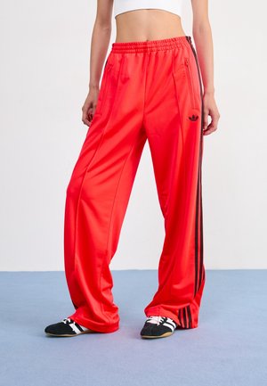 Personne portant un pantalon de survêtement Adidas rouge vif avec des bandes noires et des chaussures de sport noires sur un sol bleu clair.