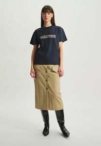 T-shirt en coton bleu marine avec un logo texte blanc et rouge, associé à une jupe en coton beige avec des boutons à l'avant et des poches plaquées. Bottes noires montantes jusqu'aux genoux.