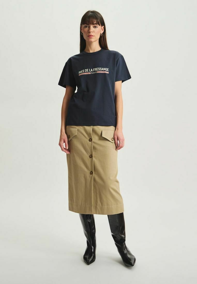 T-shirt en coton bleu marine avec un logo texte blanc et rouge, associé à une jupe en coton beige avec des boutons à l'avant et des poches plaquées. Bottes noires montantes jusqu'aux genoux.