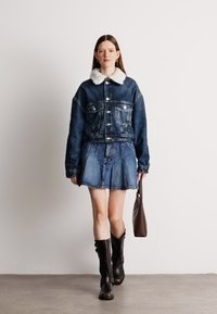 Τζάκετ από τζιν με λευκό συνθετικό γούνινο γιακά, συνδυασμένο με τζιν φούστα. Το μοντέλο φορά καφέ μπότες μέχρι το γόνατο και κρατά καφέ τσάντα.