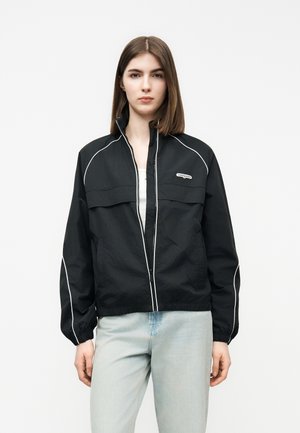 Mlada ženska, oblečena v črno jakno z belimi obrobami in svetlo modre kavbojke, stoji pred enobarvnim belim ozadjem.