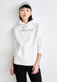 Sudadera blanca hecha de un tejido suave con un bolsillo tipo canguro, que presenta el logo "TOMMY JEANS" en negro. Combinada con pantalones negros y una gorra azul clara.