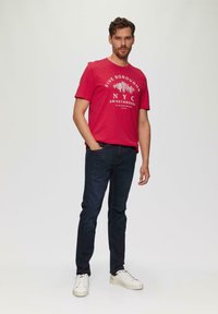 Rotes Baumwoll-T-Shirt mit dem Grafikdesign "FIVE BOROUGHS NYC DRIVETHROUGH", kombiniert mit dunklen Jeans und weißen Sneakers, steht vor einem neutralen Hintergrund.