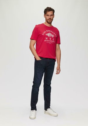 Rode katoenen t-shirt met de opdruk "FIVE BOROUGHS NYC DRIVETHROUGH", gecombineerd met donkerdenim jeans en witte sneakers, staand voor een neutrale achtergrond.